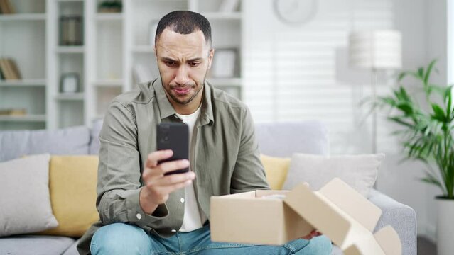 Angry dissatisfied client arabian man receive broken carton bad parcel delivery box having negative shopping experience write complaints in mobile phone give bad negative feedback at home indoor