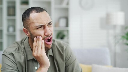 Sad handsome male suffering from toothache while sitting at home in living room. Young adult guy suffer from terrible painful tooth ache, sensitive teeth cavities Indoor in a modern interior