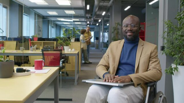 Arc portrait of male African American employee with disability sitting in wheelchair with laptop on knees in open space office smiling and looking at camera at daytime