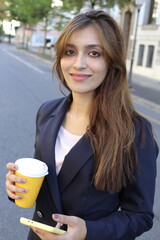 A gorgeous South Asian businesswoman walks on the street during her coffee break and smiles.