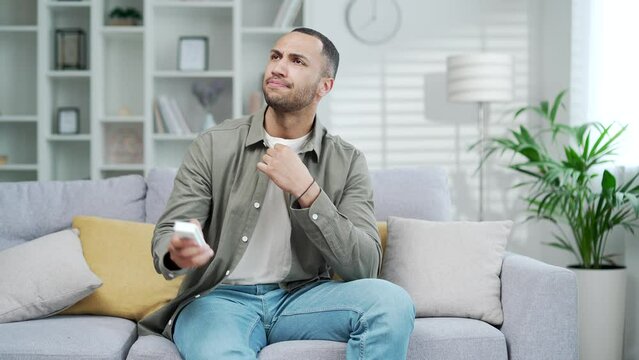 Sweaty man in hot weather is sitting at home on the sofa, he is hot and he turns on the air conditioner cooling system or humidifier Male remote control changing temperature 