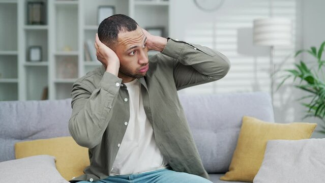 Tired man suffers because of noisy neighbors. Poor soundproofing in the apartment building and thin walls. Guy sitting in the living room and covering ears, knocking on walls indoor iside