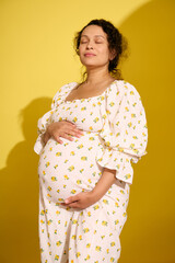 Studio shot of a beautiful curly haired pregnant woman in summer sundress, isolated over yellow studio background