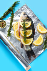 Aluminium foil roll with fresh raw fish, lemon slices, rosemary and spices on color background,...