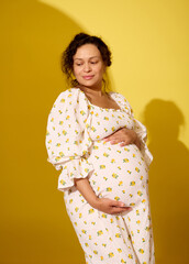 Obraz premium Studio shot of a beautiful pregnant woman in summer sundress, touching her big belly, isolated yellow studio background
