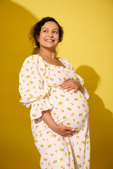 Smiling curly haired pregnant woman in summer sundress, touching her big belly, isolated over yellow studio background