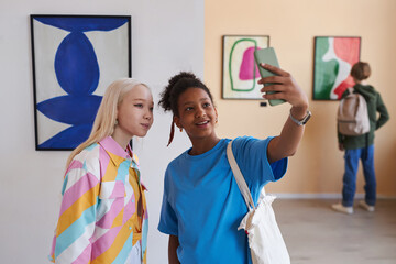 Waist up portrait of two teenage girls taking selfie photo in modern art gallery or museum, copy...