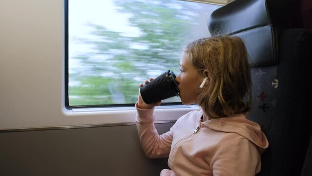 Little girl in headphones drinking hot tea or chocolate while traveling by train, looking out the window. Transportation and travel concept