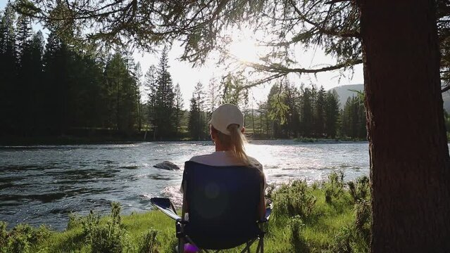 A young woman traveler is resting in the evening near a mountain river in the wild. The girl relaxes, rear view. Look at the flowing water in the river. Slow motion video. High quality Full HD footage