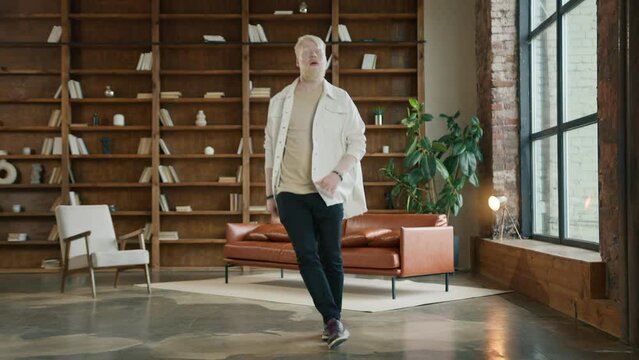 Man wearing hipster casual clothes, jumping and moving hands around in ecstatic weird moves. Blond bearded guy listening to music at home on sunny morning. Happy young man dancing in loft living room
