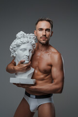 An enchanting male model, clad only in underwear, embraces a bust of a Greek sculpture, exuding charm and allure, with a gray backdrop