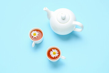 Teapot with cups of natural chamomile tea and flowers on blue background