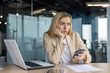 A tired, upset, disappointed business woman is sitting in the office at the desk, holding the phone in her hand and resting her head in boredom, looking to the side - Powered by Adobe