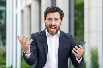 An angry man, a businessman, shouts at the camera, holds a mobile phone in his hands, spreads his hands in displeasure. Close-up photo