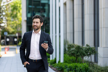 Smiling young businessman man in a business suit is using a mobile phone on the street, walking on a break outside, waiting for a meeting