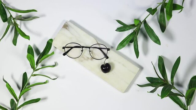 Branch with fresh green Ruscus leaves with Round Glasses Women on marble podium, and hand decorated a cherry fruits