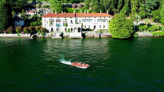 Aerial 4K footage of touristic villa Monastero located in Varenna resort, on the shore of lake Como, Italy with an old classing luxury wooden speedboat sailing and passing by