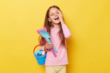 Funny laughing little girl wearing casual clothing holding sandbox toys and bucket isolated over yellow background covering half of face having fun at playground.
