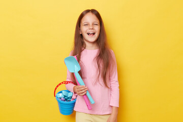 Positive happy joyful little girl wearing casual clothing holding sandbox toys and bucket isolated over yellow background laughing enjoying playing with puddle at playground.