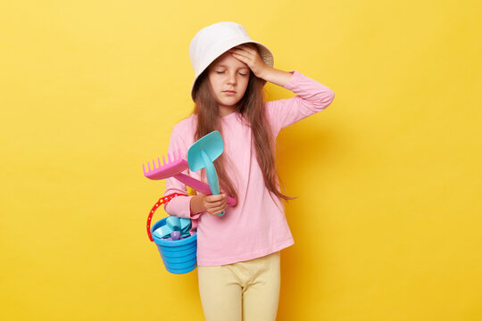 Unhealthy little girl wearing casual clothing and panama holding sandbox toys and bucket isolated over yellow background suffering headache overheated in sun.