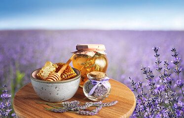 Lavender honey concept - bee honey in a jar, fresh honeycombs in a bowl and dry lavender flowers on wooden board. Rural landscape with lavender field in soft focus with farm products on display.