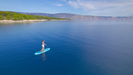 Fototapeta na wymiar AERIAL: Gorgeous shore and blue sea water with lady and her dog paddling on SUP
