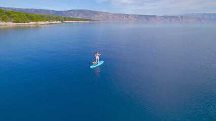 Fototapeta na wymiar AERIAL: Sporty lady paddles a SUP along picturesque coast with her dog on board