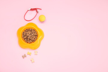 Bowl of dry pet food, collar and toy ball on pink background