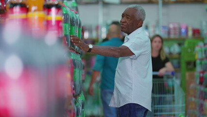 African American older shopper at supermarket searching for product to buy at soda shelf aisle