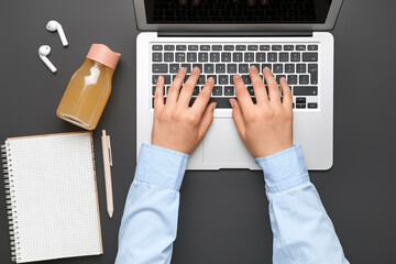 Female hands with modern laptop, earphones, notebook and bottle of juice on dark background