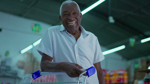 One happy Brazilian senior shopper smiling at camera with shopping cart inside supermarket shed. Portrait of an African American older consumer at grocery store
