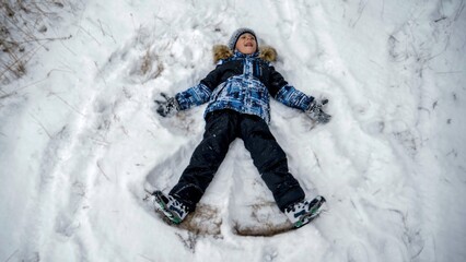 Fototapeta na wymiar Funny little boy lyin on snow and making snow angel in winter park