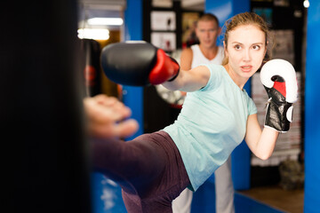 Determined athletic young woman in boxing gloves practicing heel kick by straight leg on heavy...