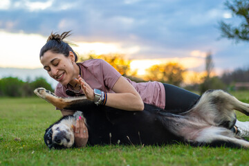Young woman playing with her dog outdoors in the backyard. Happy puppy lying on his back receiving...