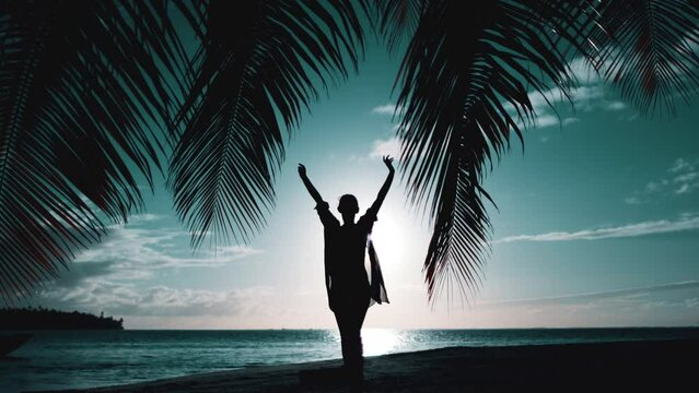 Woman walking on night tropical beach. Silhouette of slim girl under the palm coconut tree raise hands up. Bright sky with sun rays reflected in water. Blue filter toning. Slow motion. Travel, tourism