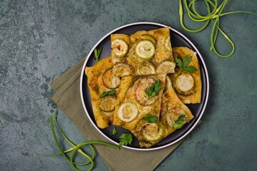 Unsweetened yeast pie with zucchini, pesto sauce and garlic arrows on a black plate on a green concrete background. Recipes for zucchini.