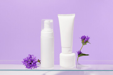 White mockup tube, jar of cream and natural foam on purple isolated background with flowers. The concept of dermatological face and body skin care products to reduce rashes. Image for your design