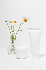 White mockup tube and jar of face and body moisturizer on white isolated background with vase of chamomile flowers. Concept of natural cosmetics for healthy skin, beauty and spa. Vertical image