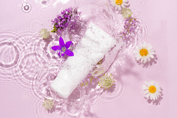 White mockup of a blank tube of cream in splashes of water on a pink background with chamomile flowers. Concept of beauty, spa, skin care
