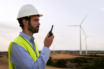 Wind turbine renewable energy engineer working and talking on walkie talkie wearing hard hat and professional safety clothing. Specialized professional people. Electric energy concept. 