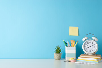 Student's workplace concept. Side view photo of white desk with alarm clock, notepads, penholder and office supplies on blue isolated background with copy-space for text or advert