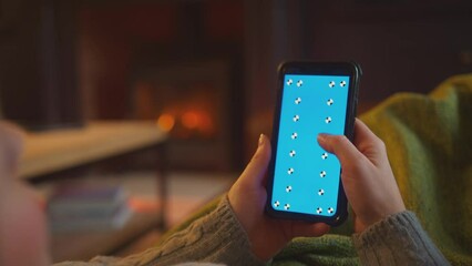Close up of woman lying on sofa with blanket at home looking at blue screen mobile phone with cosy fire in background - shot in slow motion
