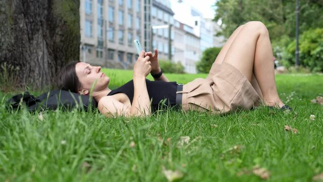 Young woman lies on a green lawn in the park on a summer day and enjoys talking on the phone. Relax in nature.