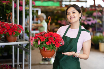 Smiling middle-aged saleswoman holding petunia in flower pot in open-air plants market