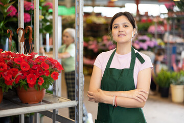Smiling middle-aged saleswoman standing next to shelves with red petunia in open-air plants market