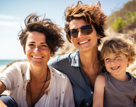 Outdoor Family Portrait Of Two Lesbian Women With Their Son.  Gay Couple And Young Boy Are Outdoors At The Beach, Looking At The Camera And Smiling, With Windblown Hair.