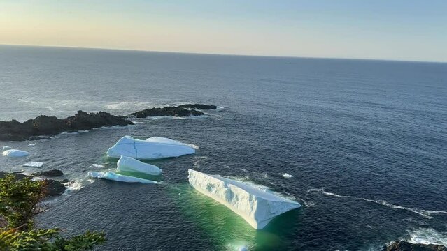 A massive tabular style or shape iceberg floating in the cold Atlantic Ocean. The large ice mass is stark white against the deep blue sea and sky. The berg has spires and a textured formation.  
