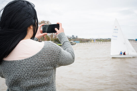 rear view of young tourist woman on the pier with her phone taking a photo of the boats on the river