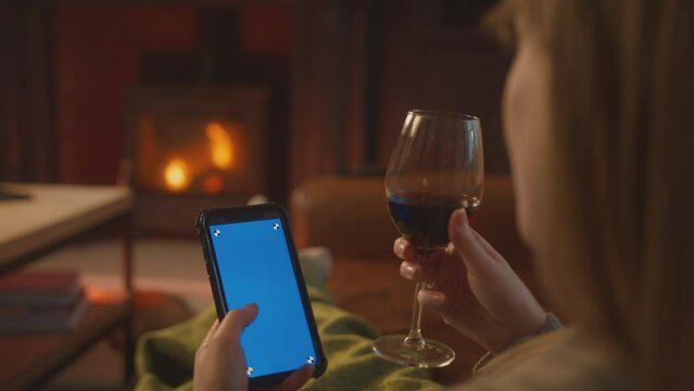 Close up of woman sitting on sofa with blanket at home looking at blue screen mobile phone drinking glass of wine with cosy fire in background - shot in slow motion