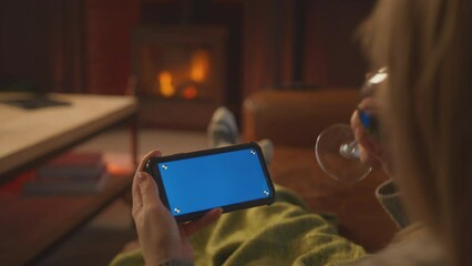 Close up of woman sitting on sofa with blanket at home streaming movie to blue screen mobile phone drinking glass of wine with cosy fire in background - shot in slow motion
